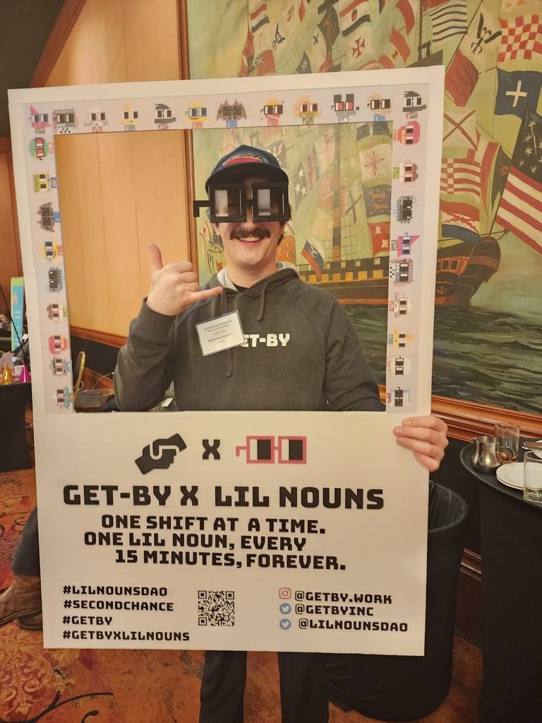 Dimitros is holding a Polaroid-style cardboard cut-out that reads "Get-By X Lil Nouns" and wearing oversize glasses. He is smiling and making a "hang loose" symbol with his hand. 
